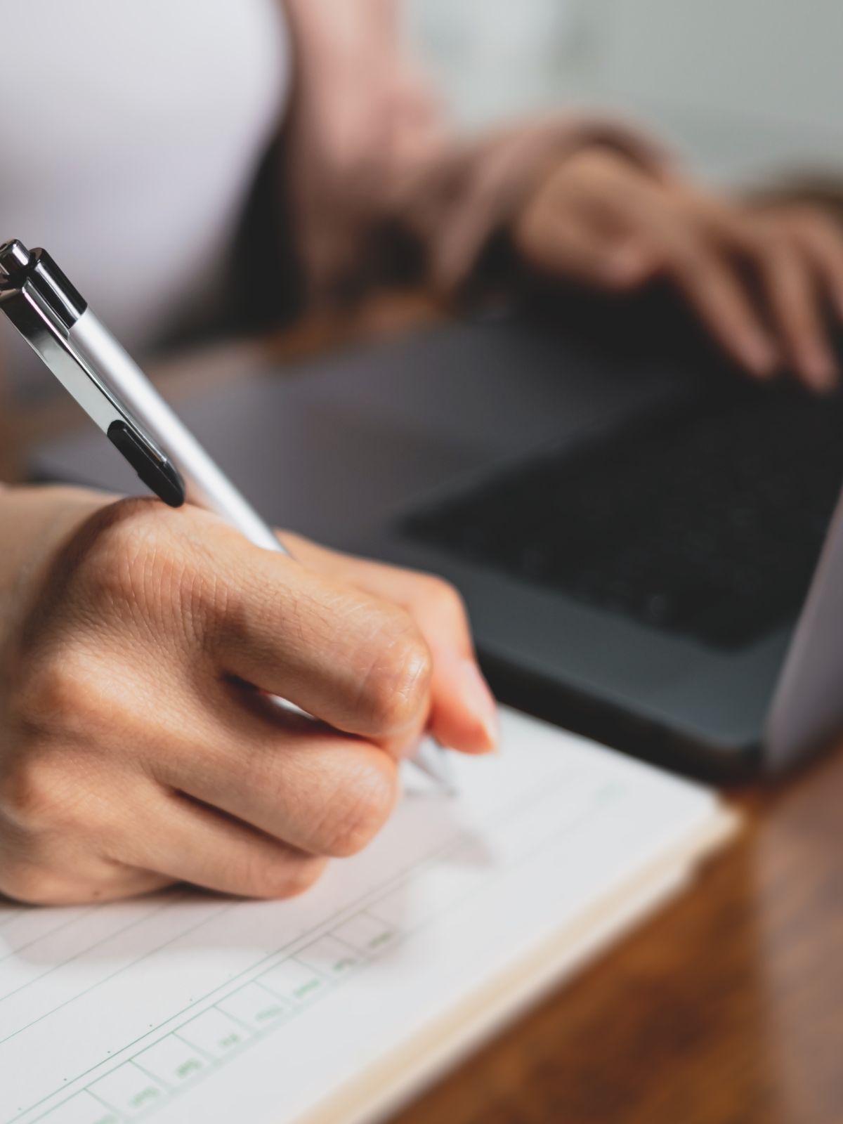 Hands writing in a notebook beside a laptop